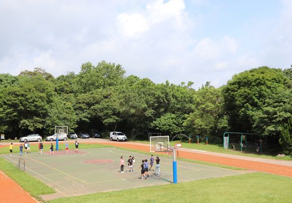 戶外運動場地，有多個籃球場和排球場。學生們在場上進行體育活動，背景為茂密的綠樹和晴朗天空。場地設施完整，包括標準的籃球架和排球網。
