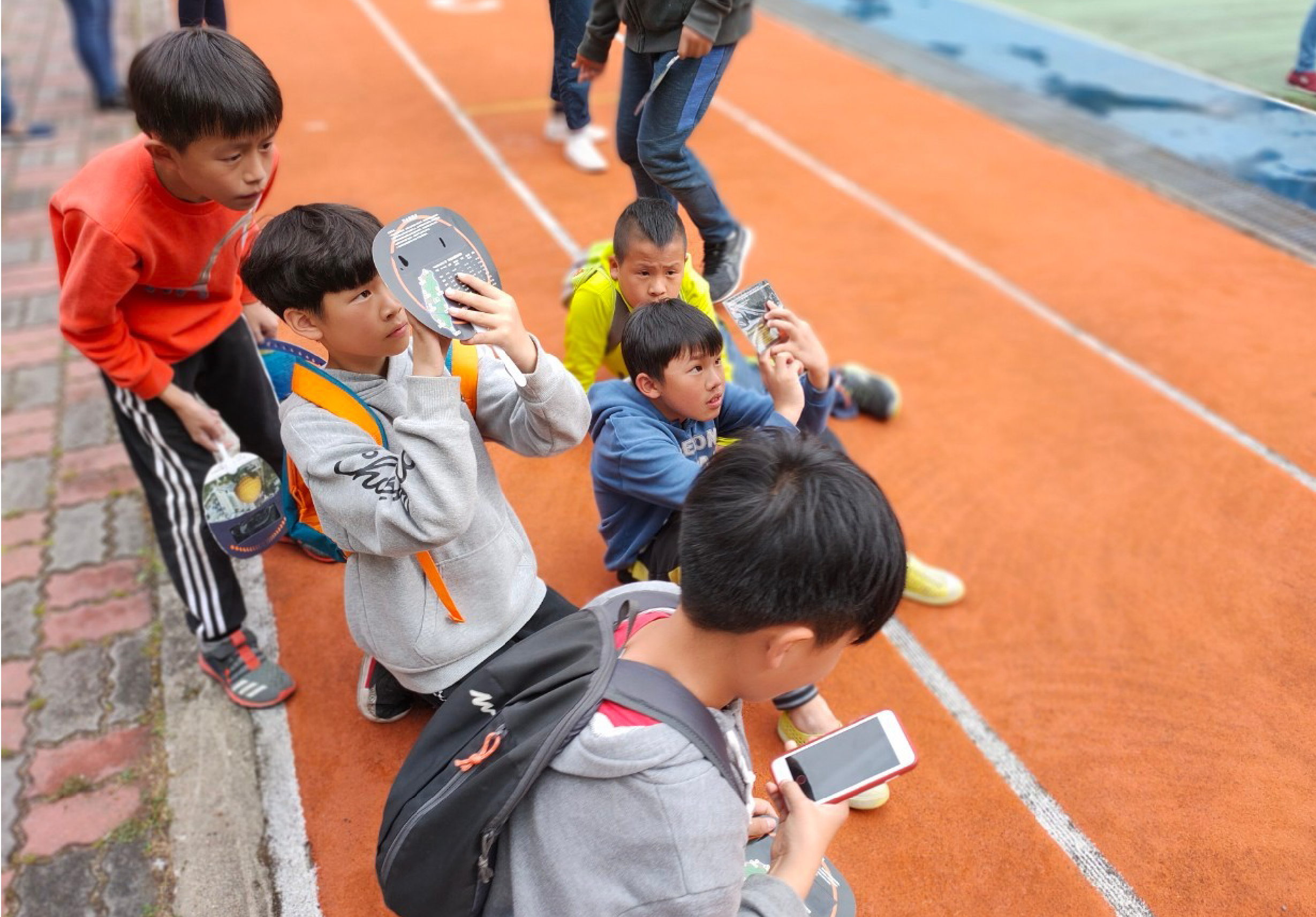 一群小學生站在橙色跑道上，幾位學生舉著手機和計步器，似乎在進行體育測量或運動追蹤活動。背景可見其他成人監督者和藍色的運動設施。