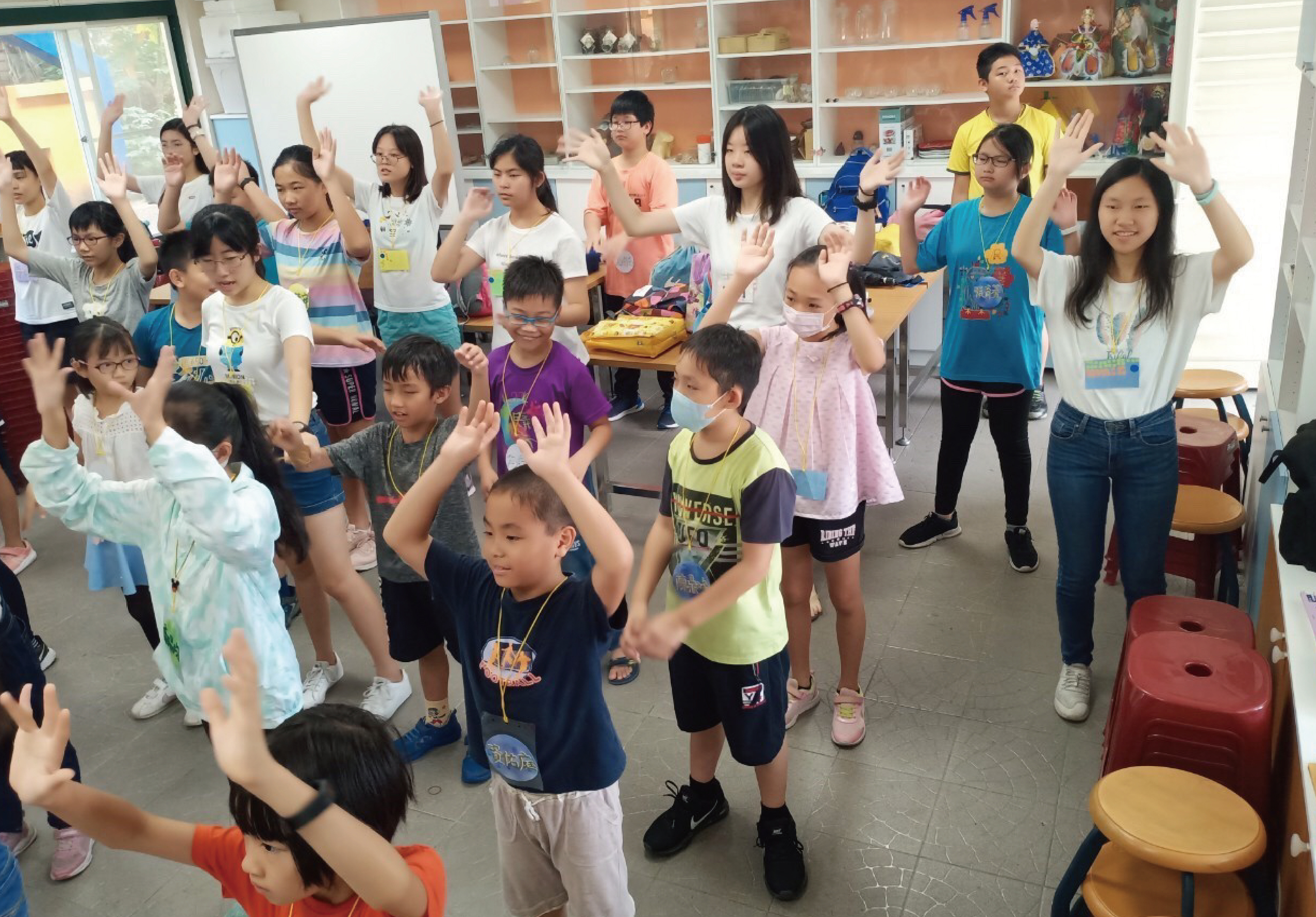 教室內一群小學生和老師進行團體活動，學生們舉起雙手，展現歡樂的學習氛圍。背景是裝滿教具的書架，地上有紅色塑膠凳子，呈現活躍的課堂互動場景。