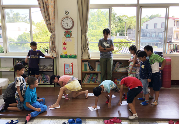 小學教室內，約十五名學童正進行體操或伸展運動。孩子們穿著校服，在木質地板上做出各種彎腰、拉伸的動作。教室背景有大窗戶、書架、時鐘和布置的牆面裝飾。