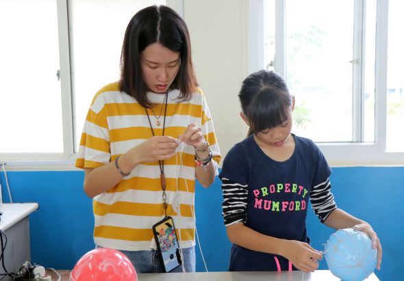 一名穿著黃白條紋上衣的女性教師正向一名穿著深藍色條紋上衣的女學生展示科學實驗器材。教室內有藍色牆面和窗戶，桌上放有紅色和藍色的球體。這是一場教育性的科學教學活動。