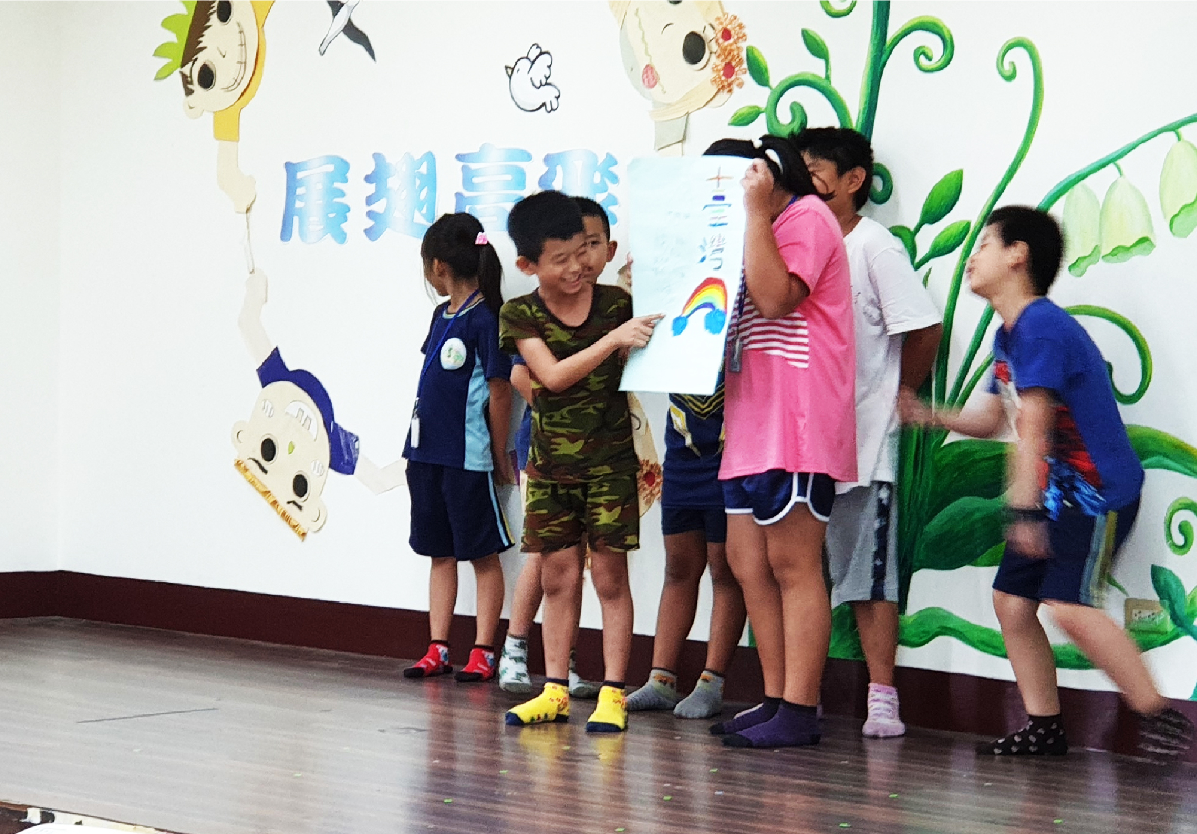 幼兒園教室內，一群小朋友圍成一圈，展示自己繪製的彩虹圖畫。牆面裝飾有卡通人物和綠色植物圖案，地板反映出溫暖的學習環境。孩子們開心地參與藝術展示活動。
