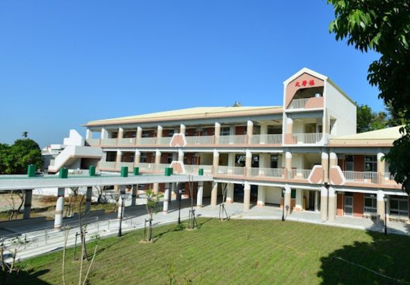 一棟現代化白色建築，三層樓高，配有多個陽台和走廊。建築周圍環繞綠色草坪和樹木，前方有步道和無障礙斜坡。晴朗藍天下，建築物設計典雅，適合作為學校或公共機構使用。