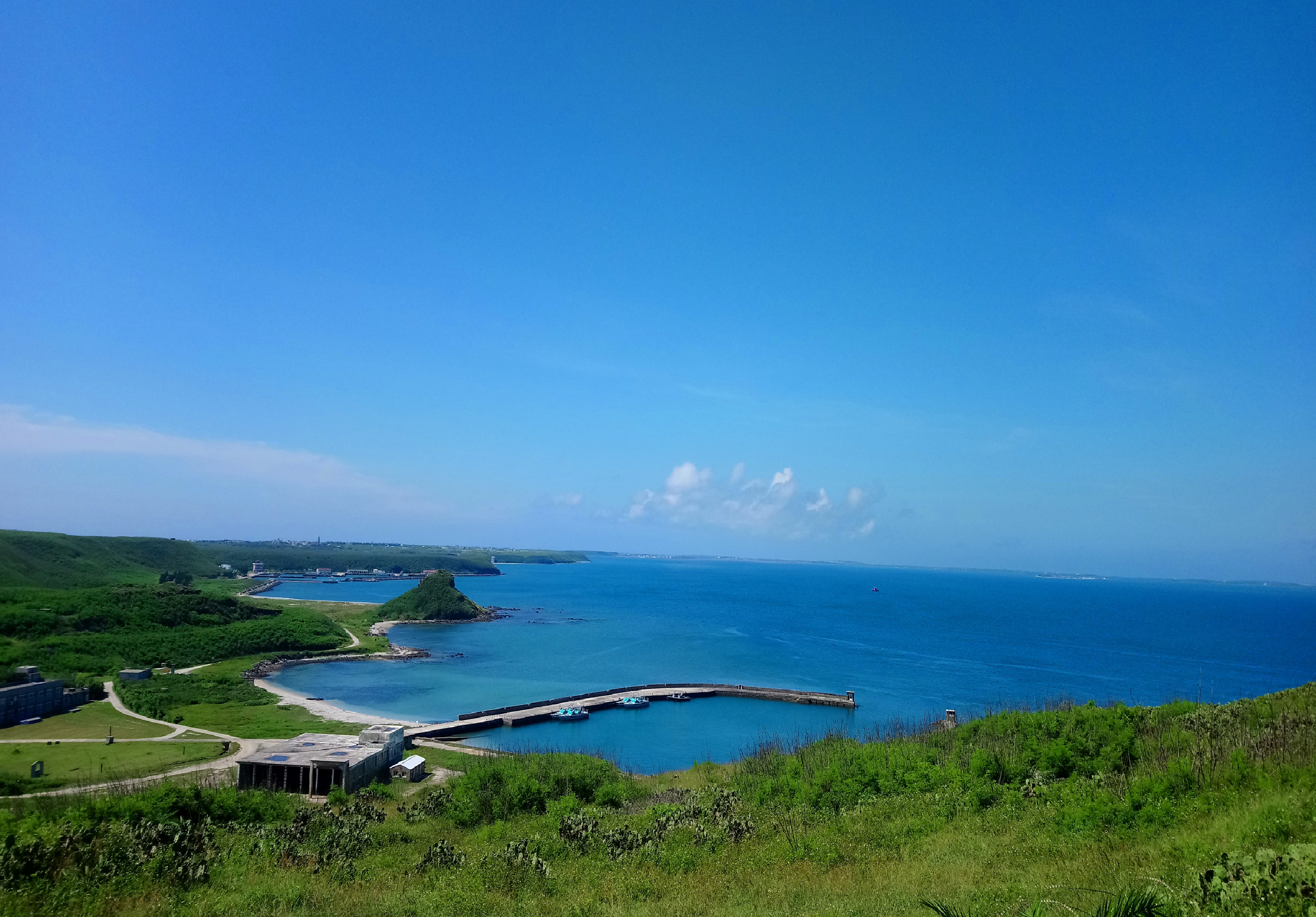 翠綠山坡俯瞰碧藍海灣，防波堤圍繞寧靜港口，遠處海面波瀾壯闊，晴空萬里，展現北東亞沿海漁村的自然風光與人文景觀。