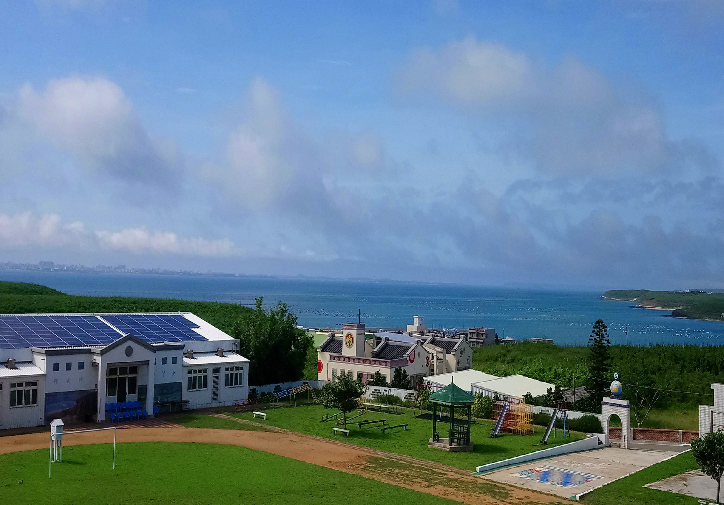海邊山坡上的校園景觀，白色建築物配備太陽能板，綠草地上有遊樂設施、沙坑和涼亭。遠景為藍色海灣和海岸線，天空飄著白雲。整體呈現現代化教育設施與自然環境的結合。