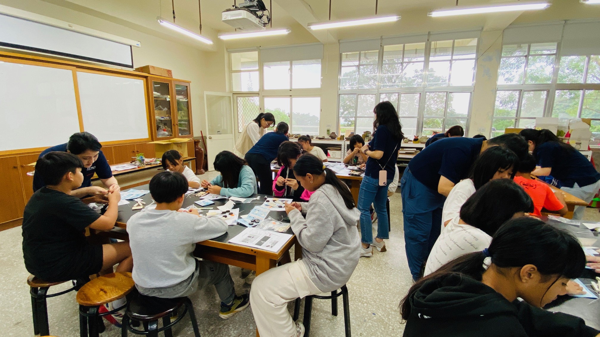 教室內進行美術課程，約二十位學生坐在木製課桌前進行繪畫或手工藝創作，數位教師在教室內指導學生。教室採光充足，配有白板、櫃子和大面積玻璃窗，可見戶外綠色景觀。