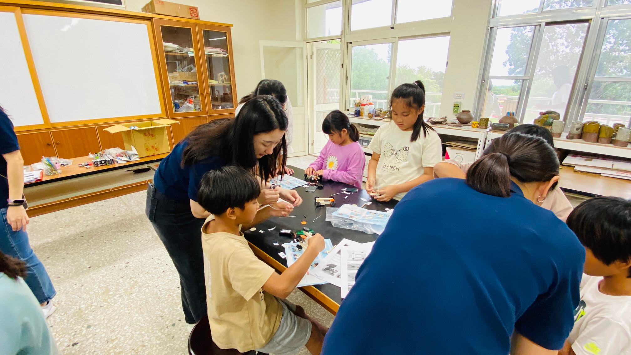 教室內，一位女性教師正在帶領多名小學生進行科學實驗活動。學生們圍坐在黑色實驗桌前，拿著教具和說明書，認真地進行操作。教室有大片窗戶採光良好，牆上掛著白板和木製櫃子。