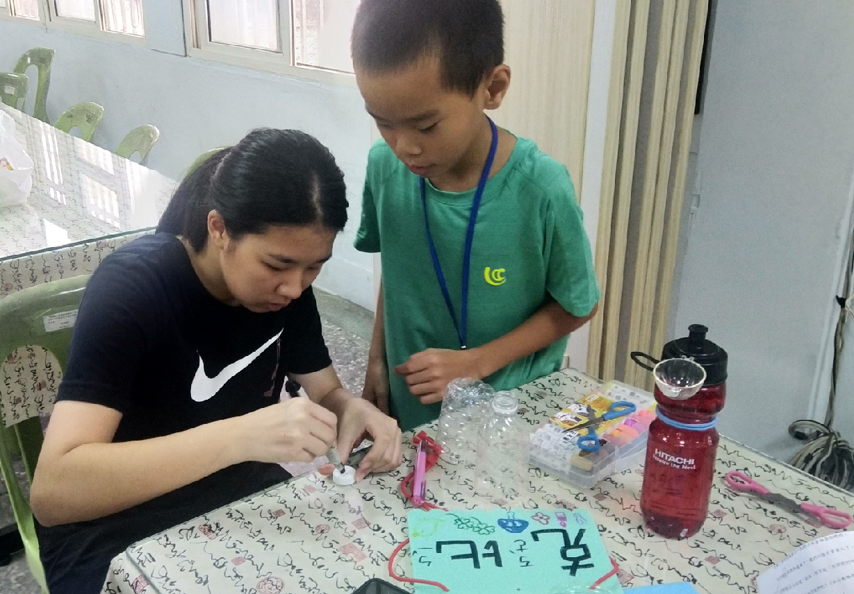 兩名學生在室內進行手工藝活動。穿著黑色Nike上衣的女學生正專注地操作彩色手工物品，穿著綠色上衣的男學生在一旁觀看協助。桌上散置著彩色工具、材料及紅色飲品，背景可見植物及窗戶。