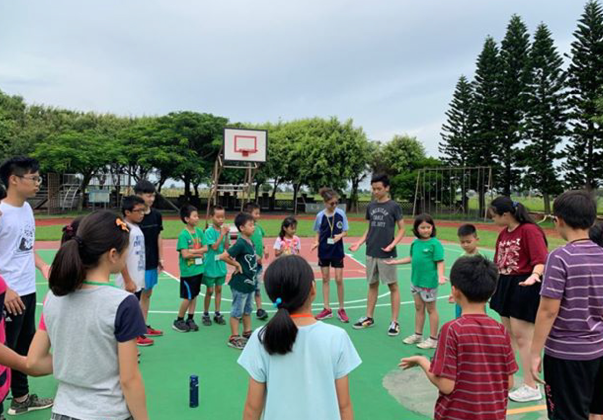 戶外籃球場上，約20多位小學生穿著各色衣服圍成一圈，參與籃球運動課程。場景背景為籃球架、綠地、樹木和晴朗天空，呈現活力充沛的戶外體育教學活動。