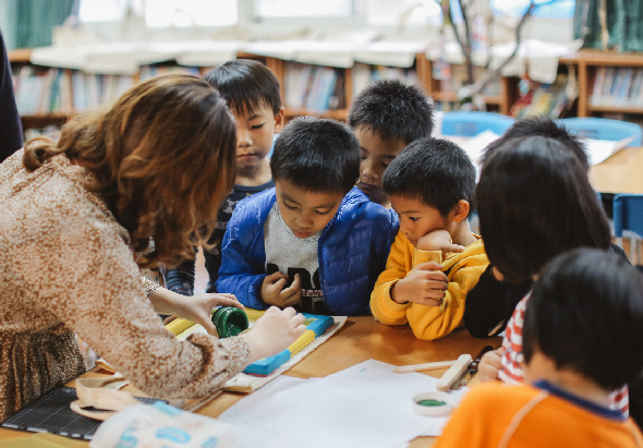 一位女性教師在教室內教導約六位小學生進行藝術創作。學生們圍坐在桌子周圍，專注地觀看教師示範手工藝品製作。背景可見書架和教室設施，呈現互動式教學情景。