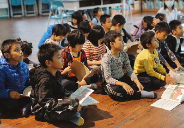 一群小學生坐在木製地板上，手持閱讀材料，專注地參與課堂活動。教室背景有藍色椅子和其他學生。這是一場集體閱讀或教學活動的現場。