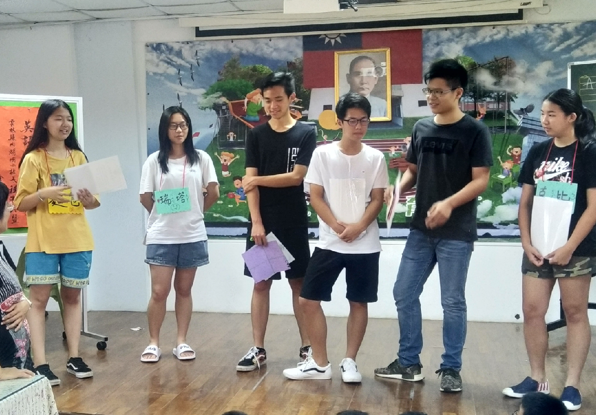 教室內六名青少年學生排成一列站立，背後牆面貼有國旗、人物肖像和彩色風景畫。左側女生穿黃色上衣，中央學生穿黑白T恤，右側女生穿花色上衣。地面為木製，天花板有投影設備。場景顯示為學校課堂或活動現場。