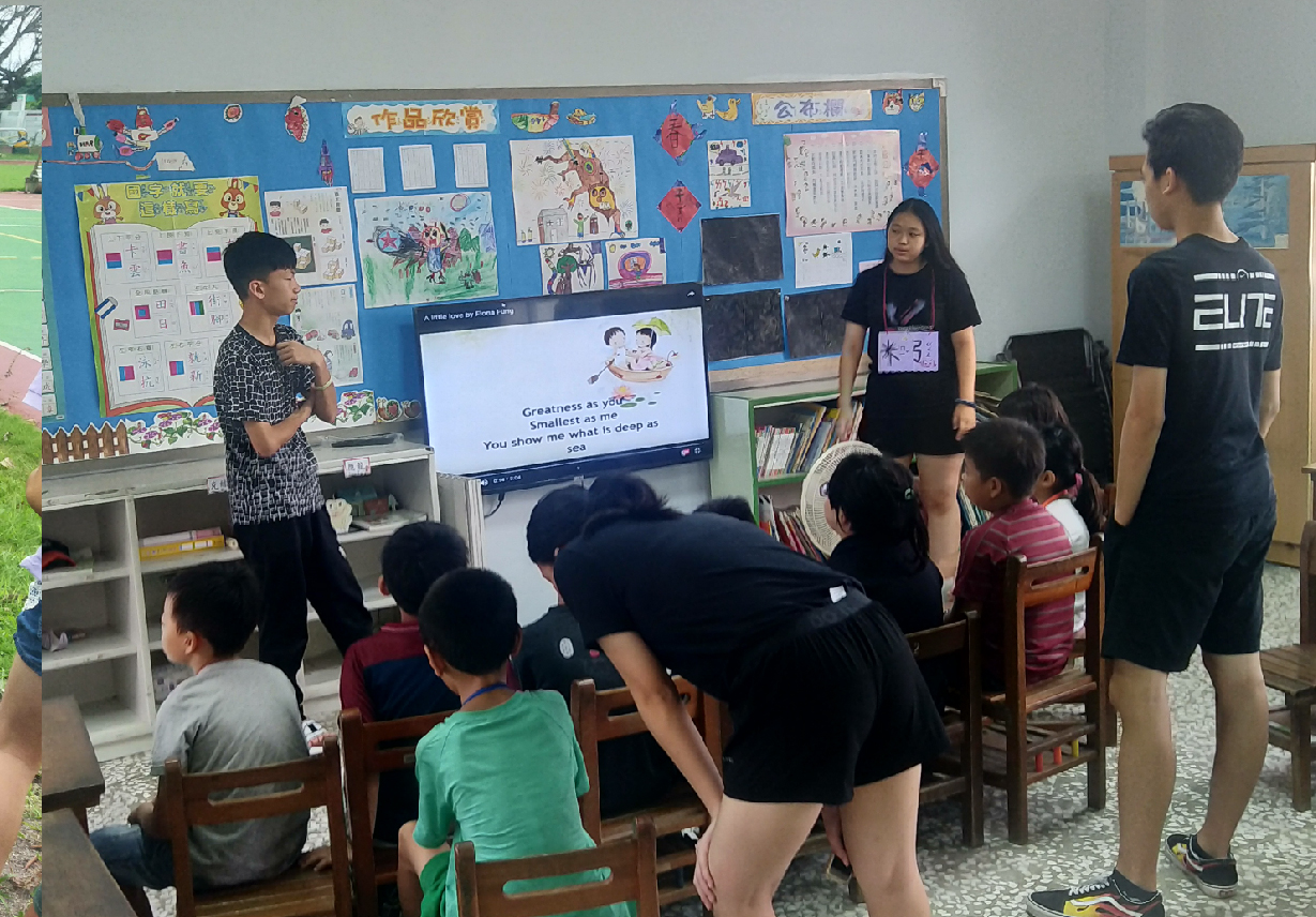 教室內，數名年輕志工用黑色衣服站立，向坐在木椅上的小學生展示投影螢幕。螢幕顯示勵志文字「Greatness at you, Smallest in size」。後方藍色佈告欄貼滿學生美術作品。這是一場教育推廣活動的場景。