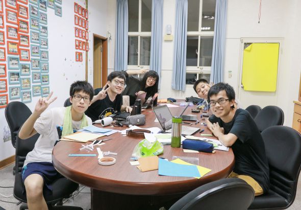 五名亞洲學生圍坐在教室的圓形棕色桌子旁，桌上擺放著筆記本、飲料和文具。牆上貼著五彩繽紛的卡片和黃色便利貼。學生們穿著休閒服裝，面帶笑容，看起來在進行小組討論或學習活動。