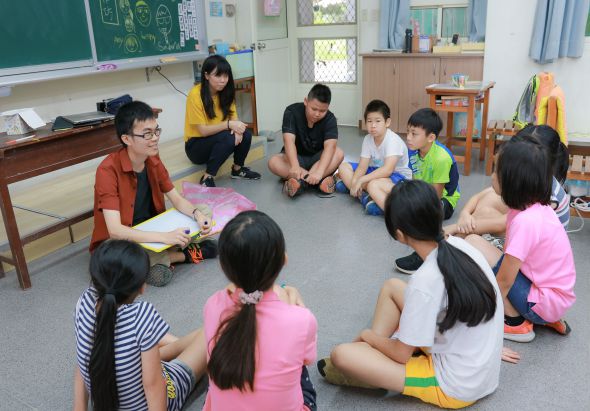 教室內，教師和學生圍坐成圈，進行互動式教學活動。學生穿著彩色衣服，教師正展示教材與學生分享，黑板和教室設施清晰可見，呈現活潑的課堂學習情景。