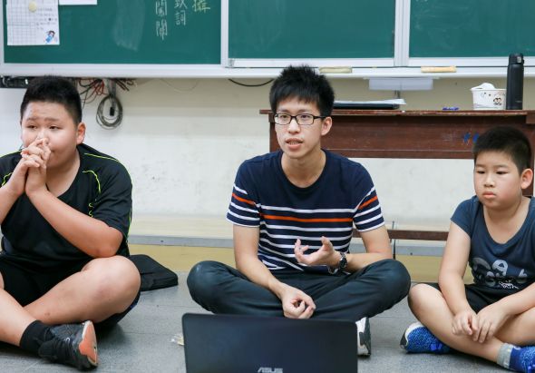 教室內三名男學生坐在地板上，中間學生戴著眼鏡，手持筆記型電腦，左邊學生托著下巴若有所思，右邊學生雙手放在膝上。身後黑板和講台清晰可見，呈現課堂學習或小組討論的情景。