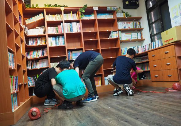 圖書館內，四名兒童蹲坐在地板上，圍成一圈進行閱讀或學習活動。身後是裝滿圖書的木製書架，牆上放著各類書籍和教材。地上有紅色球類玩具，呈現出溫暖的教育學習環境。