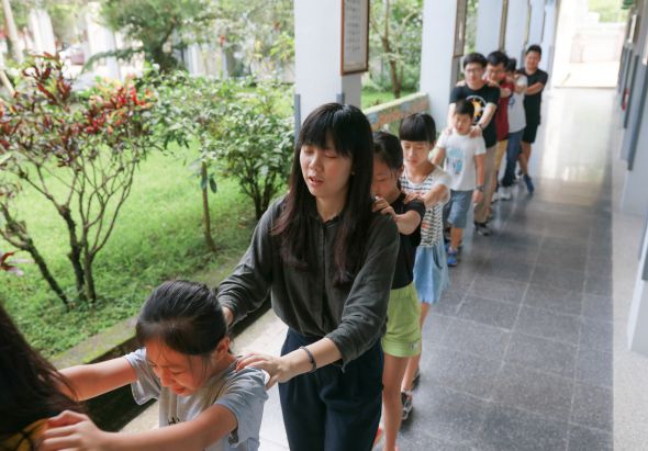 一位女性教師帶領一列兒童沿著建築物走廊行進，孩子們手牽手排成一隊。背景可見綠色草坪和植物，呈現戶外教學或集體活動的場景。