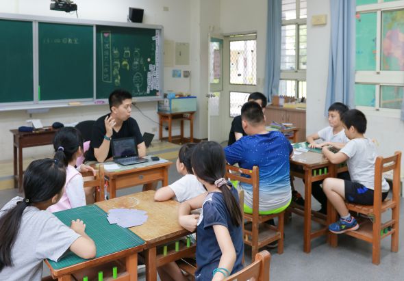 小學教室內，一位女性教師坐在學生中間，與圍坐在木製課桌旁的六名小學生進行互動教學。黑板、教室佈置和開放式窗戶清晰可見，呈現溫馨的課堂學習氛圍。