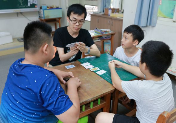 教室內五名學生圍坐在木製桌子旁進行卡牌遊戲。穿著黑色上衣戴眼鏡的學生正分配卡牌，其他學生專注地觀看綠色遊戲墊上的卡牌。背景可見教室布置和教學用具。