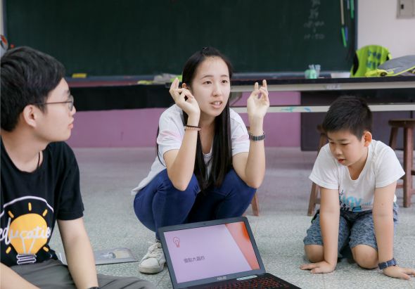 教室內，一位女性講師蹲坐在地上，面向三位學生，雙手做出手勢說明。講師身旁放有筆電，身後為綠色黑板。學生們坐在地上認真聆聽，營造互動式教學氛圍。