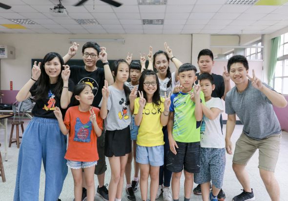 室內教室中，一群大人和小孩共約十五人排成一列，面向鏡頭做出勝利手勢，展現快樂笑容。小孩穿著彩色T恤，大人穿著休閒服裝。背景為教室設施，包括天花板、窗戶和教室用具。