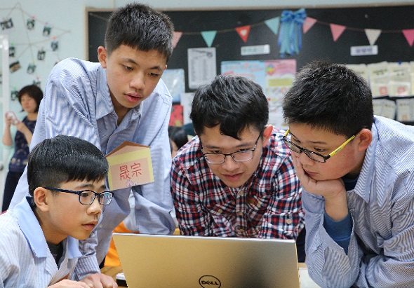 五名戴眼鏡的男學生在教室裡圍著一台Dell筆記本電腦，專注地查看螢幕內容。教室後方掛有彩色布旗裝飾，牆上貼有海報和學習資料。一名學生穿著藍色條紋襯衫站在後方，其餘四人穿著淺色或格子襯衫坐在前方。