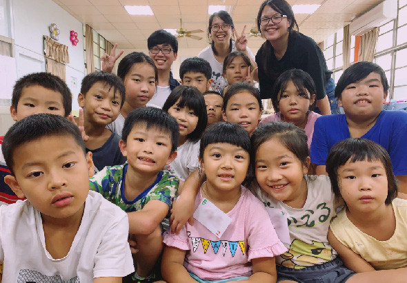 一群小學生和三位女性教師在教室內合照。學生們穿著各色衣服，笑容燦爛地聚集在一起，背景是教室裡的黑板和窗戶。這是一張溫馨的師生合照，展現校園教育現場的歡樂氛圍。