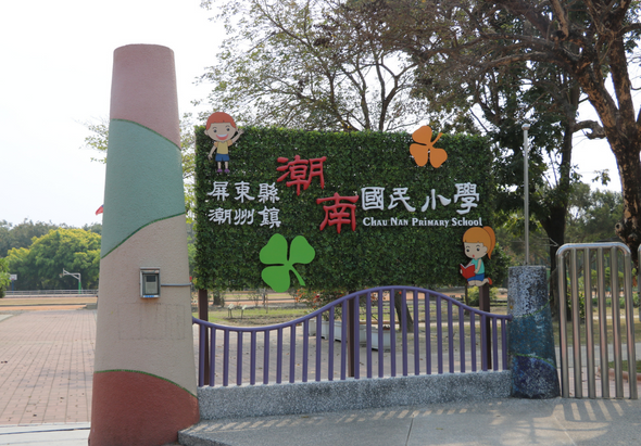 朝南國民小學校門入口，綠色籬笆牆面上有紅色校名字樣和卡通人物裝飾，左側為紅綠相間的圓柱造型，上方有大樹，鐵製校門敞開。