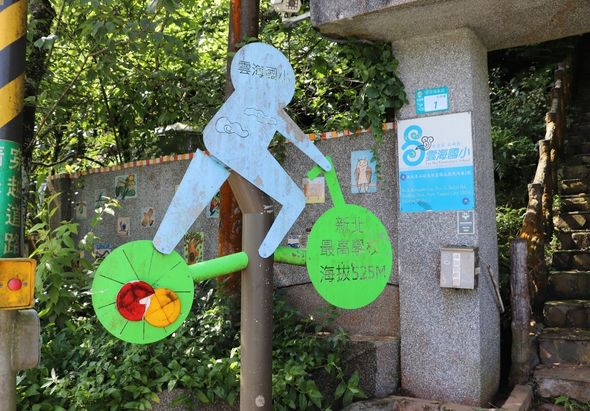 街道角落的藍色人形運動雕塑，雙手各握著一個綠色圓形物體，象徵運動健身。背景為混凝土牆面和綠色植被，旁邊豎立著藍色指示牌和公告欄。