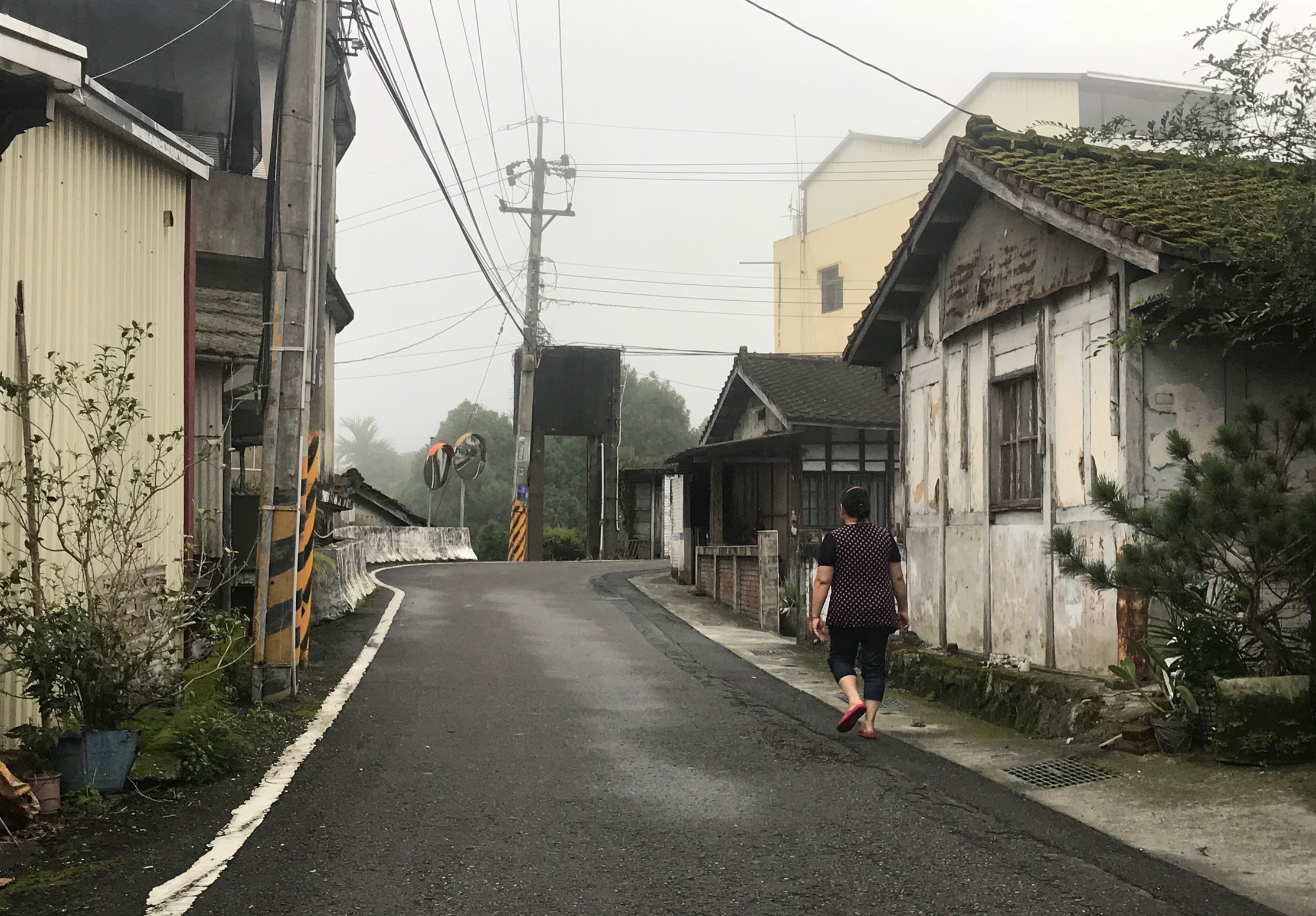 一位穿著黑色上衣的女性在濕潤的鄉村道路上慢跑。道路兩側是老舊的白色建築和傳統日式房屋，長滿了苔蘚。遠處可見高樓和電線桿，天空陰沉多雲，環境顯得寧靜而蒼涼。