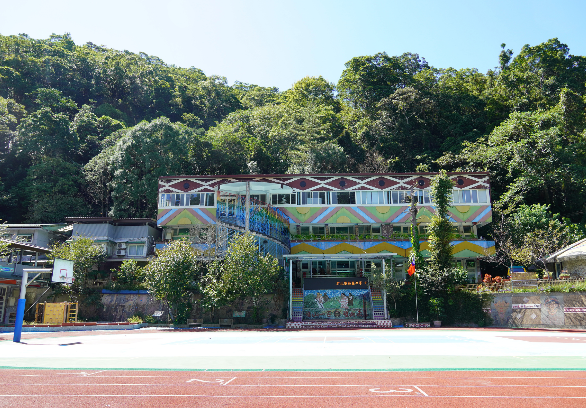 一棟彩色校舍建築坐落在翠綠山林前，外牆繪有藍色、綠色和黃色的幾何圖案裝飾。建築前方是紅色跑道和運動場，左側有籃球架。建築風格現代，展現教育機構的活力特色。