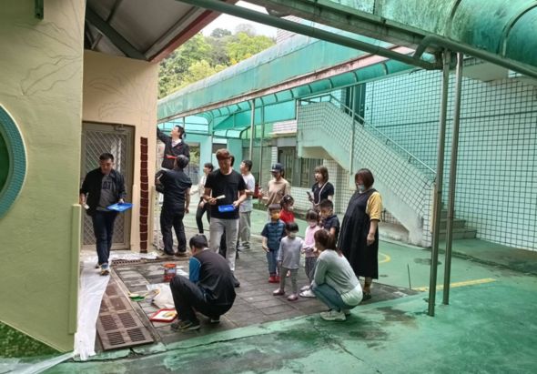一群大人和小孩在室內活動場地聚集，場地有綠色遮棚和網子。成人蹲下與幼童互動，準備進行團體活動或遊戲。背景可見教室入口和戶外綠色設施。