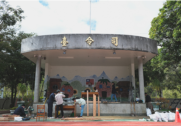 圓形舞台建築，頂部有金色中文字。舞台背景繪有彩色建築和山景壁畫。舞台前方紅色地面上有多位工作人員進行搭建或準備工作，周圍綠樹環繞，顯示為戶外公園活動場地。