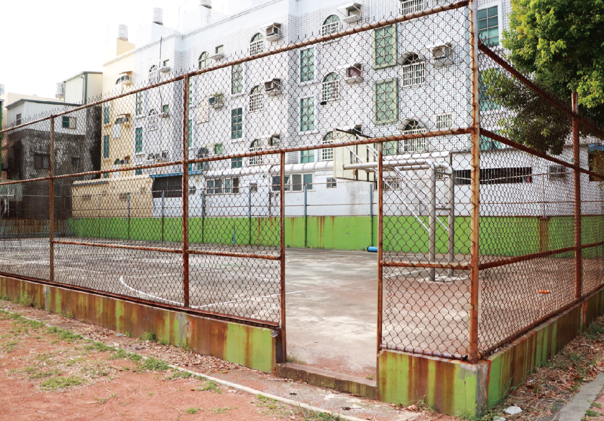 一座被生鏽鐵鍊圍籬圍繞的運動場地，內部有綠色人工草皮和混凝土地面。背景是白色磚牆的多層建築，窗戶密集排列。圍籬底部因潮濕而長滿綠苔蘚，顯示設施年久失修。