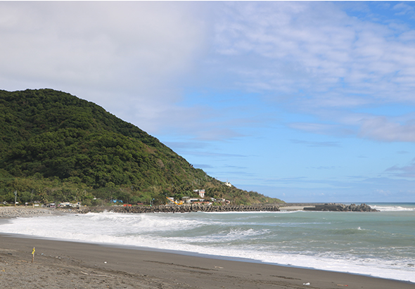 海邊沙灘景觀，左側為茂密的綠色山坡，中央是白色沙灘與翻滾的海浪，遠方可見海濱建築與住宅區，晴朗藍天白雲，海水呈現翠綠色。
