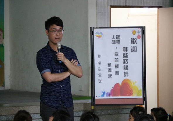 一位年輕男性講師穿著深藍色短袖襯衫，戴著眼鏡，手持麥克風在教室進行演講。身後的展示板以繁體中文撰寫歡迎詞和講師介紹，裝飾有紅黃色花卉圖案。前景可見聽眾的後腦勺，營造出課堂或講座的氛圍。