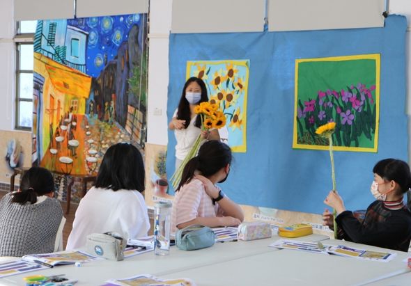 美術課堂中，一位戴口罩的老師向學生展示梵谷的向日葵作品。學生圍坐桌前觀看，身後藍色背景牆上展示著梵谷的三幅經典畫作，包括咖啡館夜景和向日葵等作品，進行藝術欣賞教學活動。