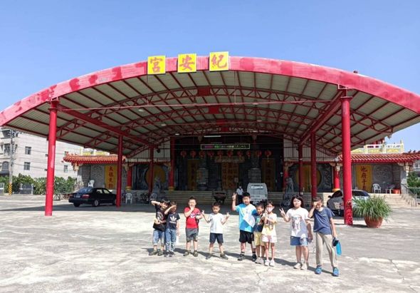 一群小學生在寺廟廣場前集合，背景為紅色弧形屋頂的建築物，頂部裝飾著黃色燈籠。晴朗天空下，孩子們身著各色衣服站成一圈，似乎進行校外教學或參訪活動。