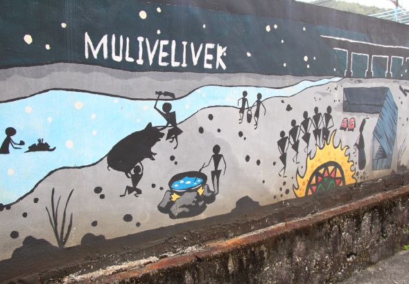 街道壁畫，上方標題「MULIVELIVEK」，描繪海灘場景。黑色剪影人物在沙灘上玩耍、游泳，有人騎摩托車、衝浪，還有黃色火焰的房屋建築。整體風格為黑白藍黃配色的現代街頭藝術作品。