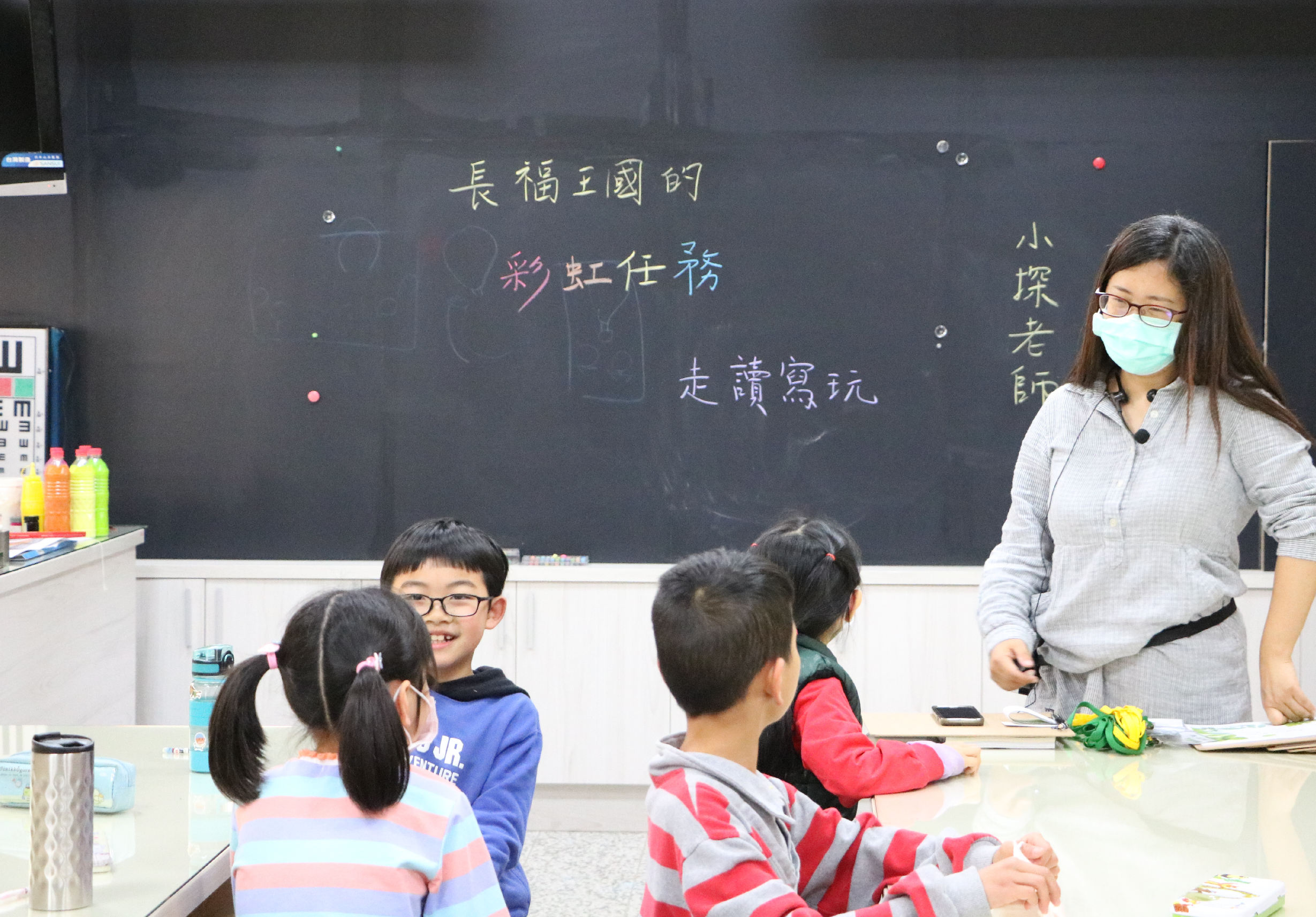 教室課堂場景，女教師戴口罩站在講台前，面向四名坐著的小學生。黑板上用粉筆寫有繁體中文，包括「長福王國的」、「彩虹住榜」、「是讀簽玩」等文字。講台左側放有教學用具和彩色筆。