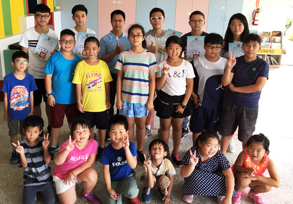 一群約二十位小學學童在教室內集合拍照，站立和蹲坐成兩排，許多學生手持獎狀或證書，背景可見布告欄和教室設施，呈現校內表揚或頒獎活動的溫馨氛圍。