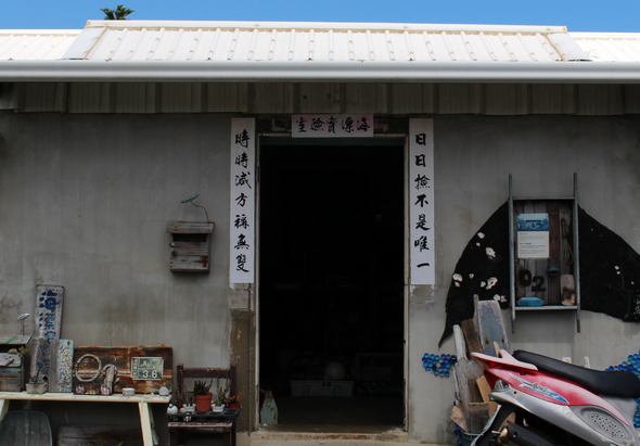 一間傳統的老屋外觀，灰白色牆面，深色木門入口上方掛著中文對聯。門兩側擺放各式物品，右側停放機車，左側窗戶開著，屋頂為白色鐵皮，整體呈現懷舊的鄉村建築風格。