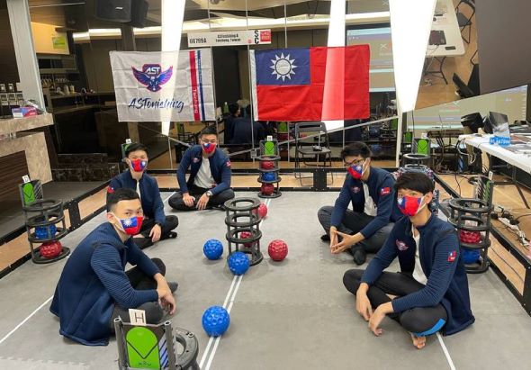 一支機器人團隊在室內場地集合，成員穿著深藍色制服圍成圓圈，中央擺放紅藍綠色球體和白色機械臂。背景懸掛台灣國旗和隊徽旗幟，展示參賽隊伍的風采。
