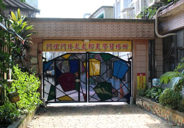 傳統建築物前的彩色玻璃門，門上方有黃底紅字的中文匾額，左右兩側種植綠色植物，地面鋪設石板，背景可見現代樓房。