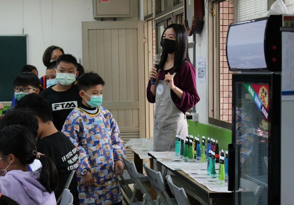 教室內，一位戴著口罩的女性講師站在講臺前用麥克風授課，面前擺放著多個綠色瓶子。約十名戴著口罩的學生圍聚聆聽，教室後方有電視螢幕和教學海報。