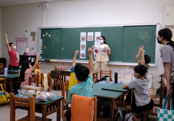 教室內，一位女教師在黑板前進行教學，數名學生坐在木製課桌椅上舉手參與課堂互動。教室布置有教學用具和學生作品，展現活躍的課堂學習氛圍。