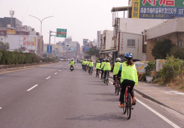 一群穿著螢光黃背心的騎士騎著自行車在寬闊的城市街道上行進，背景是日本都市建築物、廣告看板和街道標誌，天空陰沈，展現有組織的自行車隊活動。