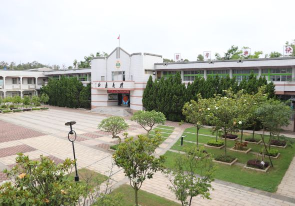 校園廣場景觀，中央為主建築物，兩側為教室樓房，廣場鋪設紅磚地面，種植多棵喬木，綠草坪整齊，路燈點綴其中，整體環境寧靜整潔。