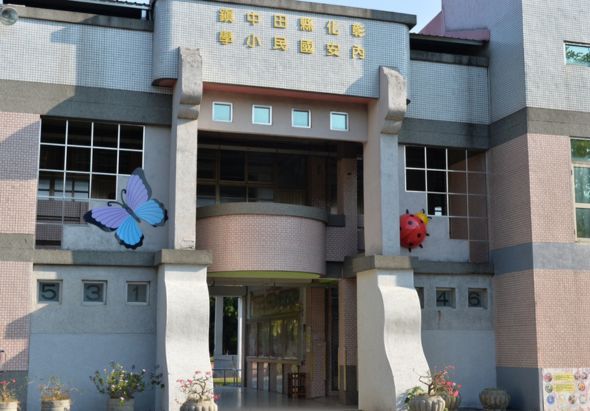 現代建築風格的小學校舍正面，米色和磚紅色外牆，中央入口上方有黃色中文校名，左側裝飾藍色蝴蝶雕塑，右側為紅色花朵裝飾，底部種植花卉，整體設計兼具教育與美感。